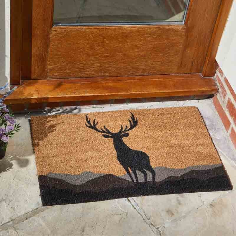 Rectangular natural coir doormat with a black stag silhouette shown in use at a wooden door.