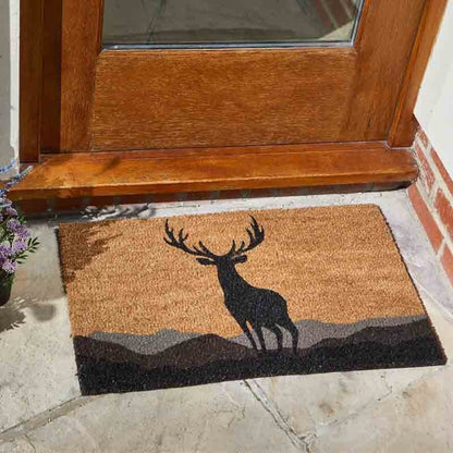Rectangular natural coir doormat with a black stag silhouette shown in use at a wooden door.