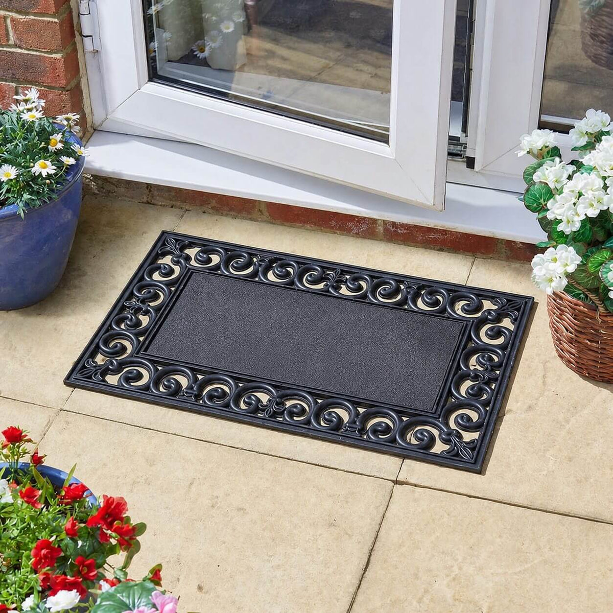 Black rubber doormat frame with scrollwork border outside a patio door, with potted flowers