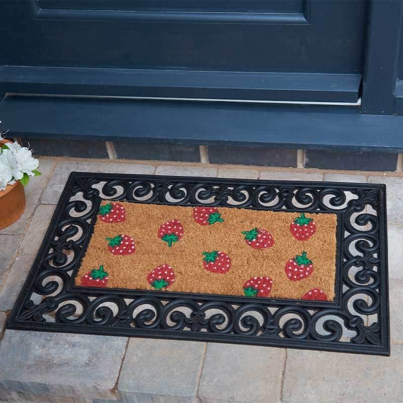 A rectangular doormat with a black scrollwork border and a tan center featuring a repeating strawberry pattern, placed on a stone step in front of a dark door.