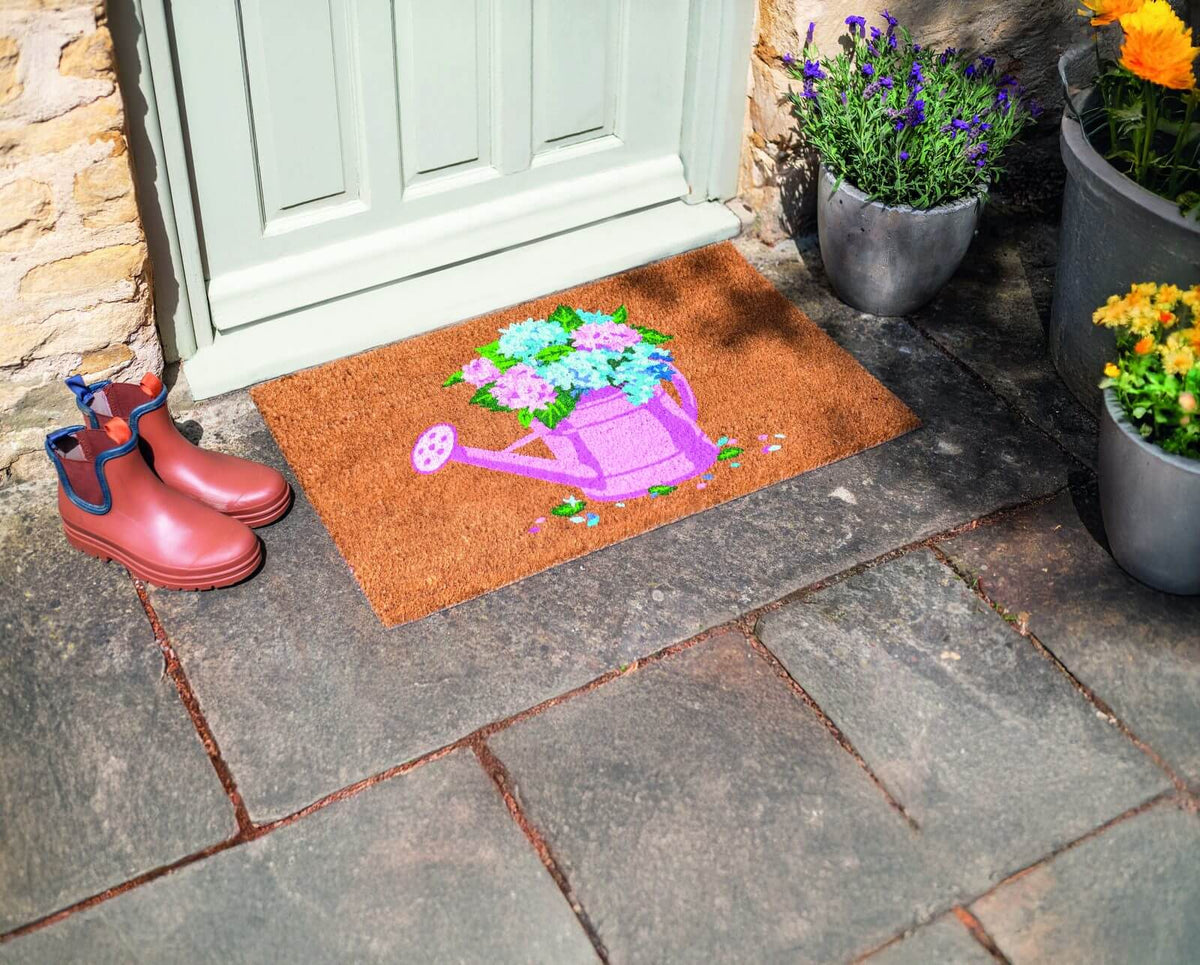 Watering bloom coir doormat with pink watering can and floral design on a stone step by a front door, with wellington boots