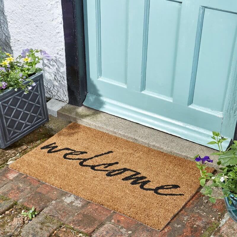 Welcome coir doormat with black script lettering on a brick doorstep by a teal front door